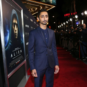 Riz Ahmed in sharp navy suit, with alien movie poster in background. Red carpet event