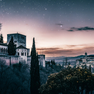 Dusk falls over Granada, casting a mix of purple, orange, and blue hues on the historic Alhambra. City lights begin to twinkle.