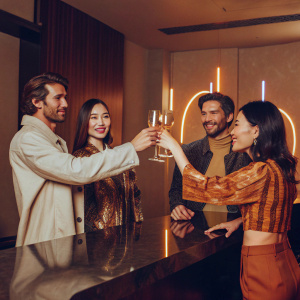 David Beckham, Kiko Mizuhara, Angelababy, and Adrianne Ho celebrate at a sleek, modern bar. Neon lights flicker behind them, casting vibrant colors across their faces. They clink champagne glasses in a joyful toast.
