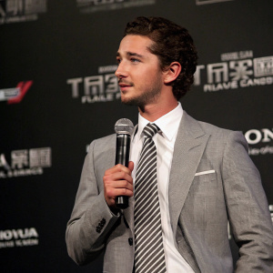 Shia LaBeouf, in a gray suit with a white shirt and striped tie, holding a microphone. Dark backdrop with company logos. Event setting, press conference style.