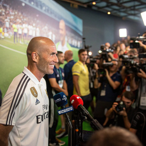 Zinedine Zidane, the bald head coach of Real Madrid, during a vibrant stadium press conference. He's wearing the club's iconic all-white jersey.