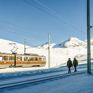 A serene winter scene at Rotenboden station in Zermatt, Switzerland. A brown tram stands still, framed against a backdrop of snow-covered mountains. Dark-clad pedestrians dot the snowy landscape.