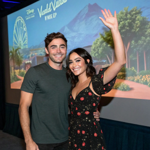 Zac Efron and Vanessa Hudgens attending a virtual fan meet-up. Zac in a dark grey t-shirt, Vanessa in a black floral pattern dress. The backdrop is a digital screen showing a theme park landscape.