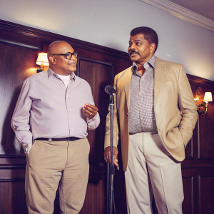 Larry Wilmore and Neil deGrasse Tyson at the Comedy Cellar. Dark wood paneling backdrop, with spotlights.