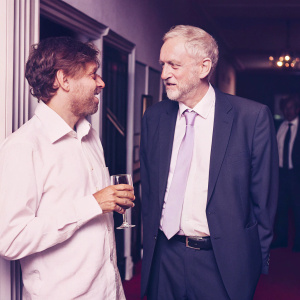 Jeremy Corbyn, with a distinguished beard and warm smile, alongside Jez Bond in a stylish dark suit and a striking purple tie. The scene captures them on the vibrant red carpet at the Park Theatre Gala 2015.