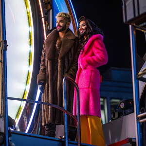 Bill Kaulitz and Conchita Wurst, attending a high-profile awards show. Bill in a brown fur coat and dark pants, Conchita in a pink, fluffy ensemble. Against a bright white backdrop.
