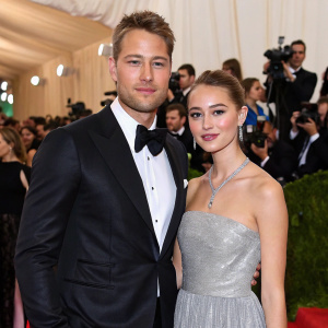 Justin Hartley and Isabella Justice Hartley at a bustling gala event. Justin wears a black tuxedo, Isabella is in a stunning silver gown. Crowds of photographers and guests in the background.