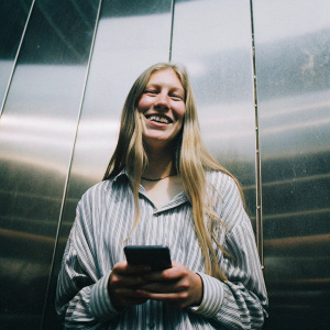 Long-haired blonde woman, casual striped shirt, smiling widely while holding smartphone. Dark, glossy background. Studio photo