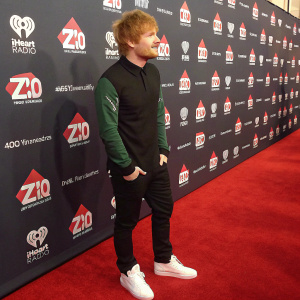Ed Sheeran at a Z100 event. Wearing black shirt with green sleeves and white sneakers. Red carpet and logo-filled backdrop