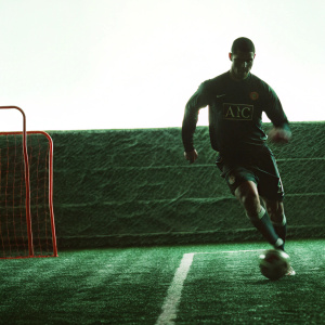 Cristiano Ronaldo, in dynamic motion on a pristine soccer field. He sports a dark Manchester United jersey adorned with the AIG logo and number 10. The backdrop showcases a well-tended green pitch and a distinctive red training net.