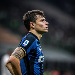 Nicolo Barella, intense focus, dark hair, tattoo on arm. Wearing Inter Milan jersey, standing on soccer field, background blurred.