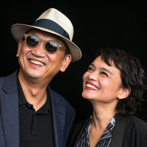 Wong Kar Wai and Norah Jones, smiling joyfully at a film festival. Wong wears a stylish fedora hat. Solid black background