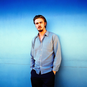Professional headshot of Boyd Holbrook, Caucasian actor. Neatly styled hair and beard. Gray shirt, black pants. Soft lighting, plain gray wall background