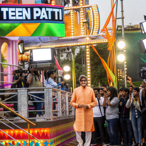 Amitabh Bachchan in a vibrant orange sherwani, promoting 'Teen Patti' at a bustling outdoor event. Surrounded by fans and media.