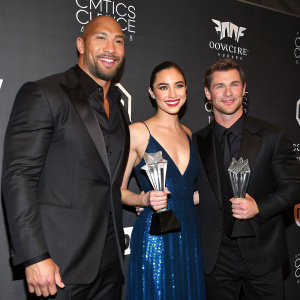 Celebratory pose at Critics' Choice Movie Awards. Dwayne Johnson, Gal Gadot, and Chris Hemsworth. They wear formal attire, dark suits, and hold trophies. Behind them, a black backdrop with award show logos.