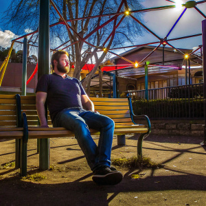 Photograph of Liam Hemsworth sitting casually on a park bench, dressed in a navy t-shirt and jeans. Sunny day with a few clouds
