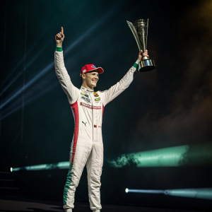 Mick Schumacher, in celebratory pose with racing trophy. Dark background with dramatic lighting.