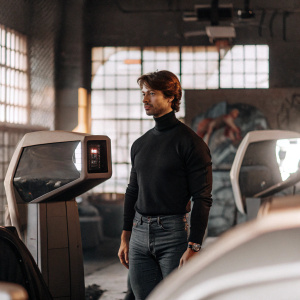 Patrick Schwarzenegger at a tech startup launch. Wearing a black turtleneck and jeans. He's surrounded by futuristic gadgets, neon signs, and urban graffiti backdrop.