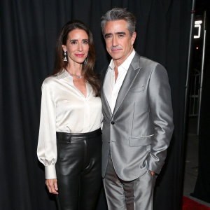 Tharita Catulle and Dermot Mulroney, stylishly dressed, attending the film festival opening night. Tharita in a white blouse paired with black pants. Dermot in a tailored silver-grey suit. Black backdrop behind.