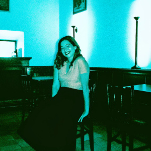 Dominika Paleta, cheerful and charming, wearing a taupe blouse and black skirt, posing for a promotional shoot in a cozy cafe setting with warm, ambient lighting.