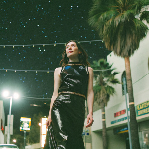 Astrid Berges-Frisbey, Caucasian actress, in a shiny black dress with gold and blue accents. Attends Hollywood movie premiere. Background is a starlit sky with palm trees.