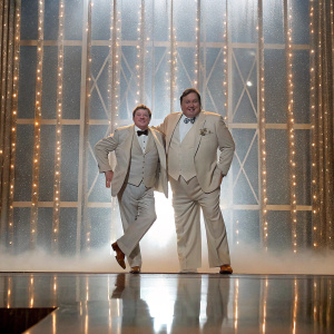 Nathan Lane and John Goodman, dressed in suits, at a Broadway award show. Glittering stage backdrop