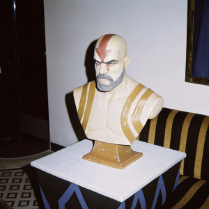 High-definition bust of Kratos, crafted in hyper-realistic skin-like material, detailed with wrinkles and facial lines, set against a white platform adorned with abstract shapes and a sleek modern background.