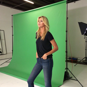 Kelly Rohrbach, a fair-skinned blonde actress, in a casual black top and jeans. Standing in front of a large green screen studio.