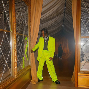 Travis Scott, posing dramatically at the Met Gala. He sports dreadlocks and a vibrant, neon-colored outfit.