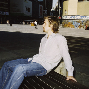 Ryan Gosling, wearing a casual shirt and jeans. Sitting on a bench in a bustling city square. Graffiti covered walls in the background.