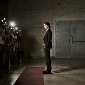 Cole Sprouse at an award show, dressed in a black suit, slicked-back hair. Red carpet with flashing cameras and paparazzi.