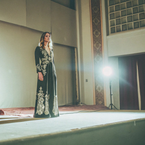 Kristen Stewart, wearing a stunning black satin dress with silver embroidery, standing alone on a dimly lit stage. No photographers in sight, only a single spotlight highlighting her