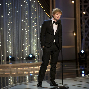 Ed Sheeran, dressed in a black tuxedo, posing elegantly on the stage of a glamorous award show.