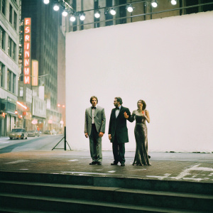 Jake Gyllenhaal, Michael Longhurst, and Ruth Wilson, standing on a stage during the opening night of a play. They are posing in front of a backdrop that displays the bright lights of Broadway.