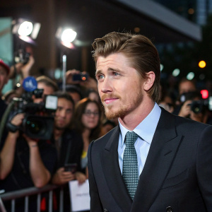 Garrett Hedlund at a press conference. He is wearing a dark suit, light shirt, and patterned tie. The background is a sea of flashing cameras and reporters.