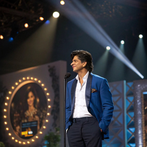 Shah Rukh Khan, in a blue blazer and white shirt, at an awards ceremony. Spotlight on him, stage behind