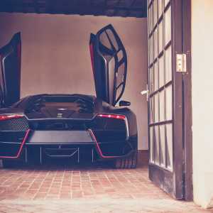 A photo of a Lamborghini Veneno parked in an open garage. Dark surroundings highlight the car's striking black body, accented with red and blue details. The sleek, aggressive design and aerodynamic features are prominently displayed.