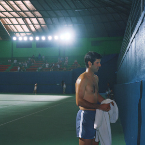 Novak Djokovic, shirtless, standing by his locker room. Drying himself with a towel. Brightly lit indoor stadium, spectators in background.