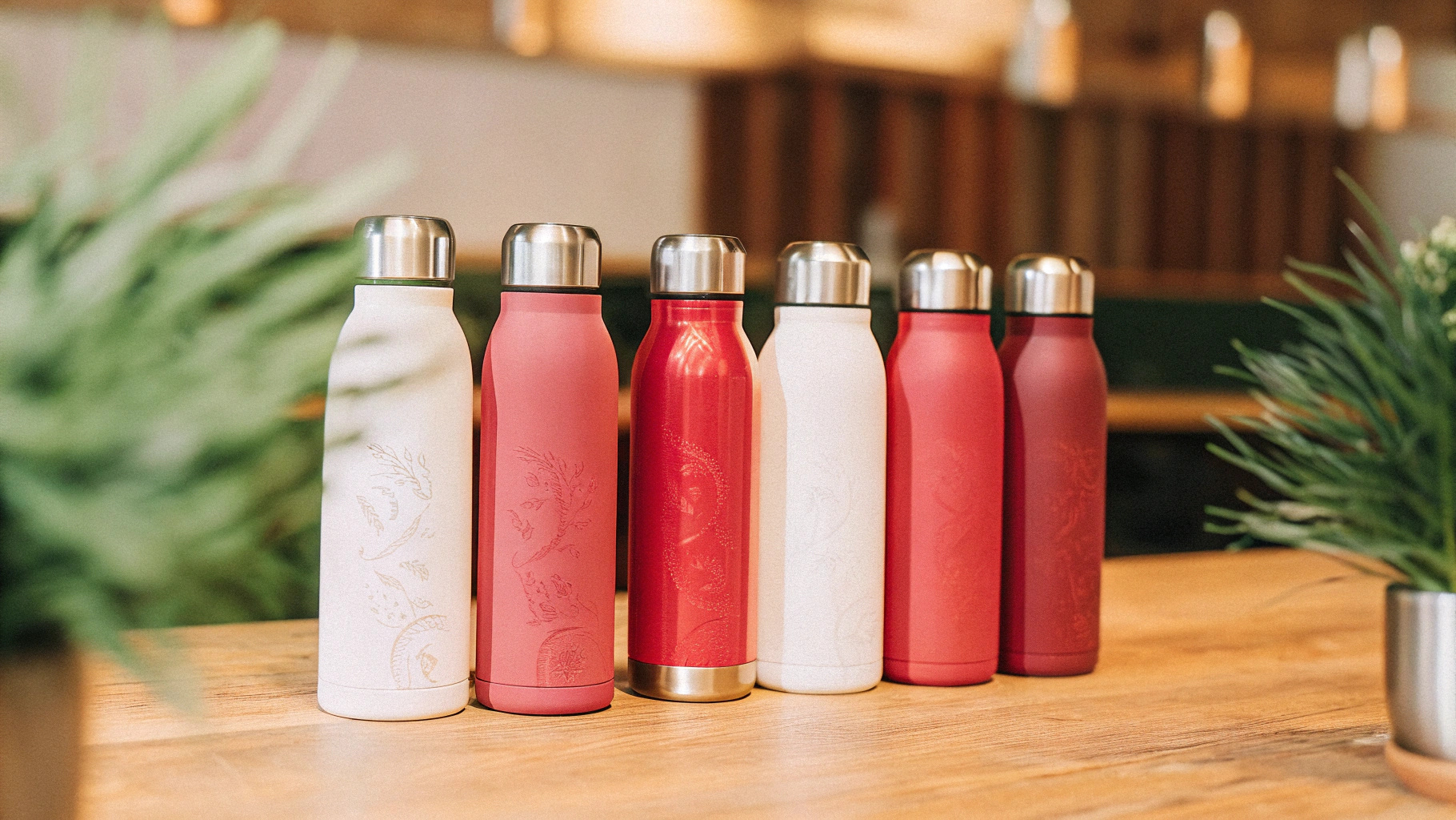 A collection of customized stainless steel insulated bottles with various colors and engraved logos, placed on a wooden table with a blurred cafe background.