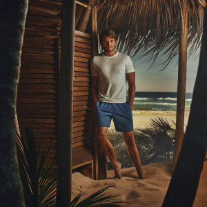 Zac Efron, posing outdoors at a casual beach setting. Wearing a white t-shirt and blue shorts. Sandy backdrop