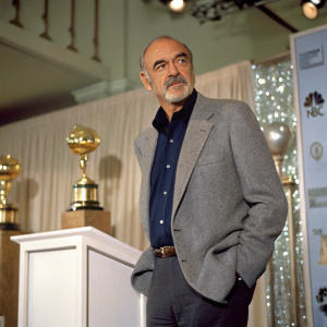 Sean Connery, in formal attire, at the Golden Globe Awards. Gray blazer, dark blue shirt, standing beside a podium.