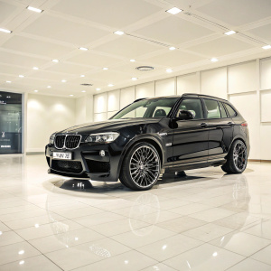Hamann's BMW X3 in a glossy showroom. Sleek, black exterior contrasts with the pristine, off-white floor. Sporty design with chrome accents on the wheels and grille. Professional photo showcasing the vehicle's refined and powerful appearance.