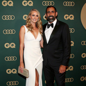 Natalie Croker and Adam Goodes at the GQ Australia Men of the Year Awards. Natalie, blonde wavy hair, white V-neck dress, high slit, beige accessories. Adam, South Asian, full beard, formal black tuxedo. Against a backdrop with the Audi logo and a rich, dark background.