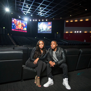 Angela Simmons and Bow Wow sit on a sleek black couch in an empty theater. Large digital screen displaying vibrant visuals behind them.
