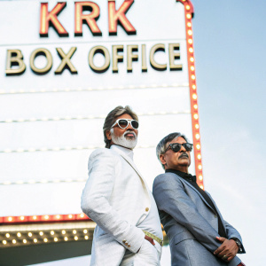 Amitabh Bachchan and Kamaal Rashid Khan at a glamorous movie premiere. They stand confidently in front of a vibrant, neon-lit backdrop featuring KRK Box Office branding.
