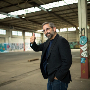 Steve Carell, wearing a dark blazer and beard, giving a thumbs up in front of an empty warehouse with graffiti.