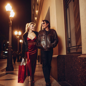 Courtney Stodden and Doug Hutchison, walking down a bustling city street at night. Courtney wears a red velvet gown and a leather jacket, holding a red shopping bag. Urban background with streetlights and passing pedestrians.