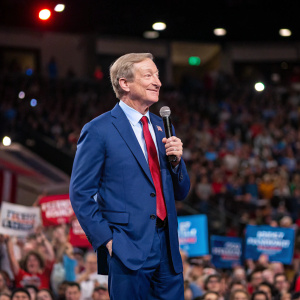 Tom Steyer, wearing a blue suit and red tie, addressing a rally. Large crowd behind him