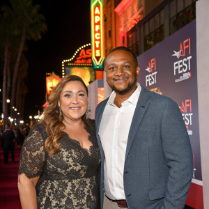 Jo Frost and Darrin Jackson, at a Hollywood event, posing in front of vibrant AFI Fest backdrop. Jo in elegant lace dress, Darrin in crisp white shirt and gray jacket.