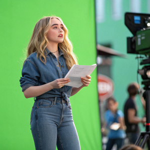 Sabrina Carpenter on set, vibrant green screen backdrop. Wearing a casual denim shirt and jeans, holding script.